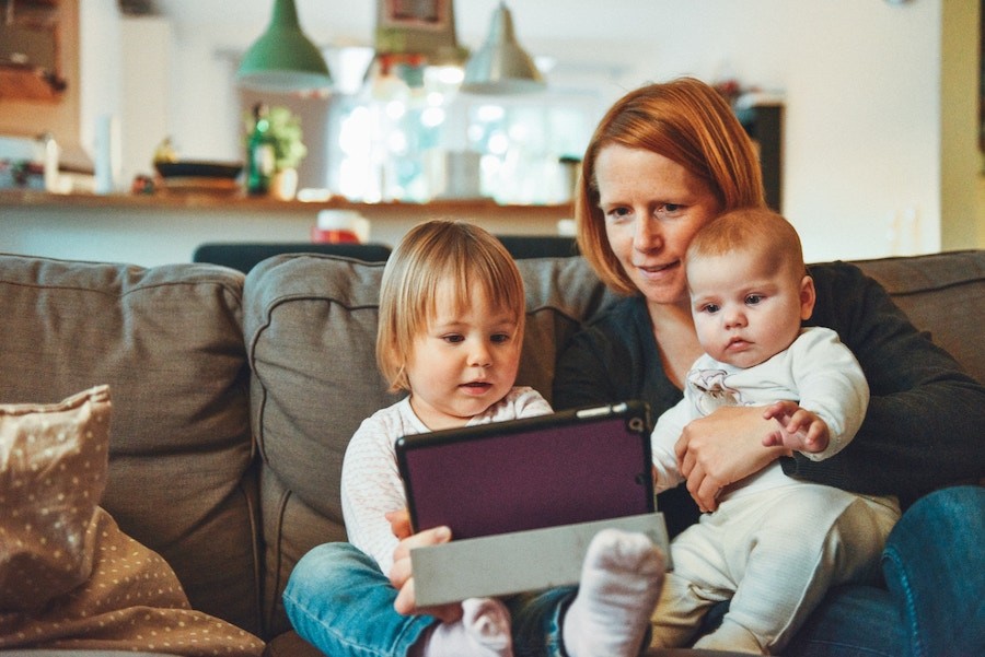 woman sits on sofa with two smaller children while looking at a tablet together