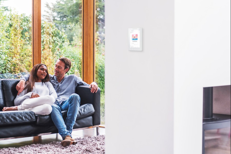A man and a woman sitting on a couch next to a wall-mounted Crestron touchpad.